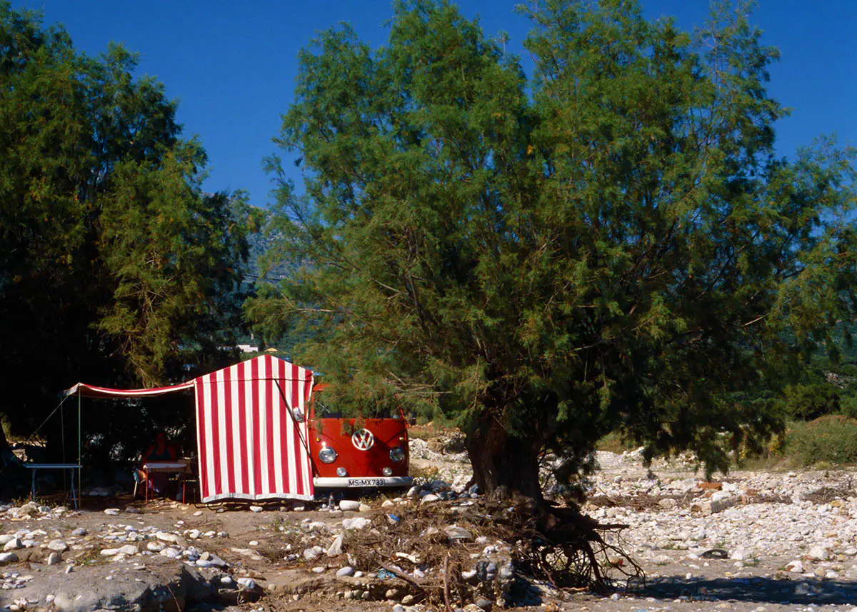 Roter VW T1 mit rot-weiß gestreiftem Vorzelt an einem naturbelassenen Stellplatz unter Bäumen